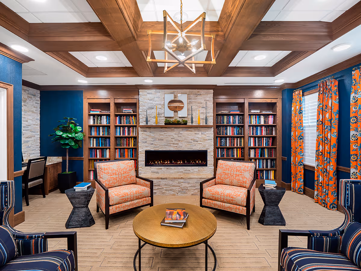 A cozy living room area with a modern stone fireplace in the center, flanked by built-in wooden bookshelves filled with books. Two orange patterned armchairs face a round wooden coffee table with books on it. Two blue patterned armchairs are partially visible in the foreground. The room features a wooden coffered ceiling with a geometric chandelier, blue walls with wooden trim, and orange floral curtains on the windows. A small desk and chair are visible to the left, along with a potted plant.