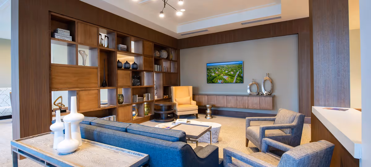 A modern lounge with blue sofas and armchairs facing a wall-mounted TV, wooden shelving and decorative accents in a communal living area.