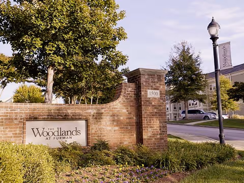 Brick entrance sign reading "The Woodlands at Furman" at a landscaped driveway entrance with trees, a lamp post, and the building visible in the background.