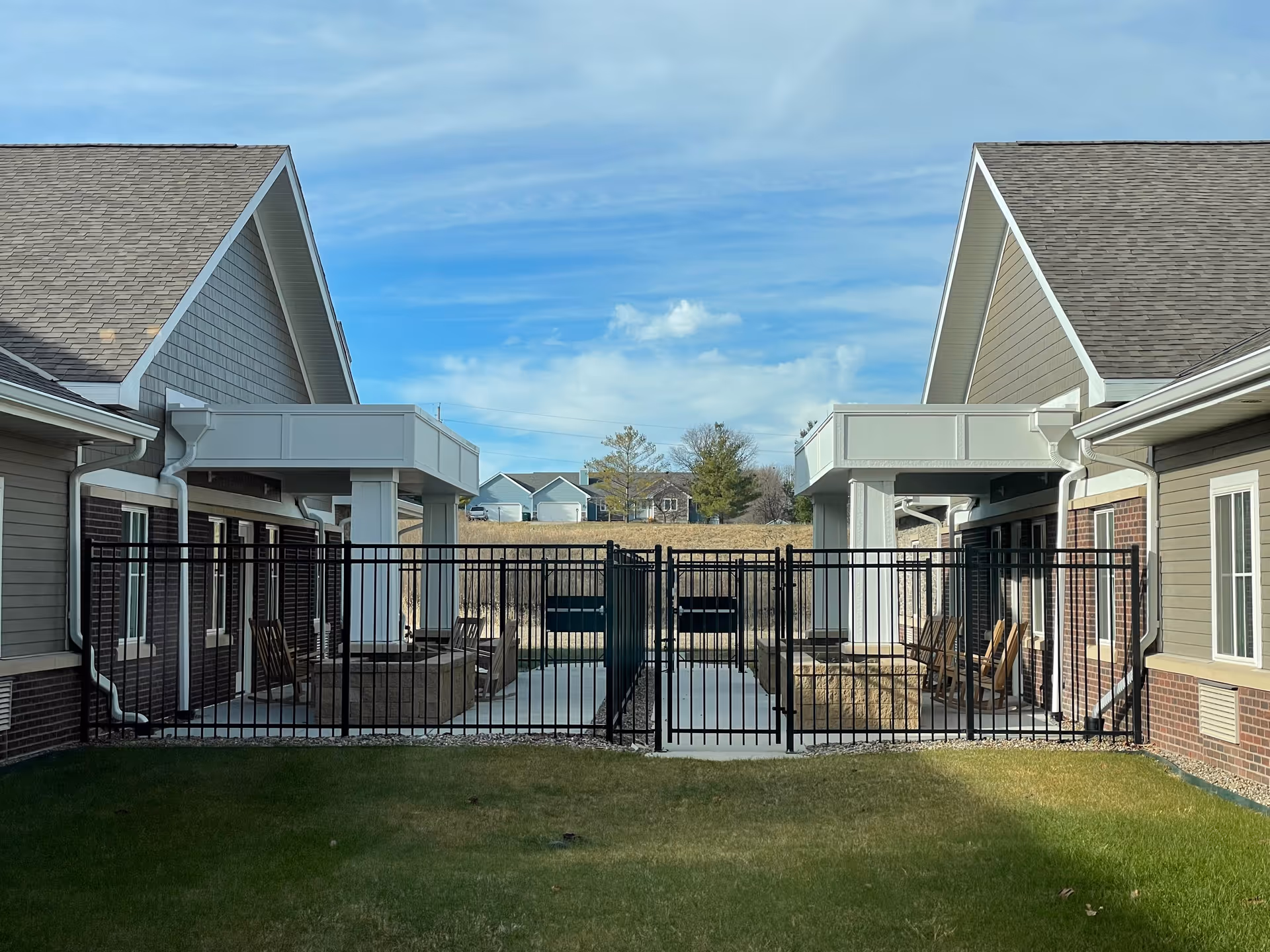 Outdoor view of a fenced patio area between two buildings with pitched roofs. The patio has seating and a fire pit, with a grassy lawn in the foreground and houses visible in the background under a partly cloudy sky.