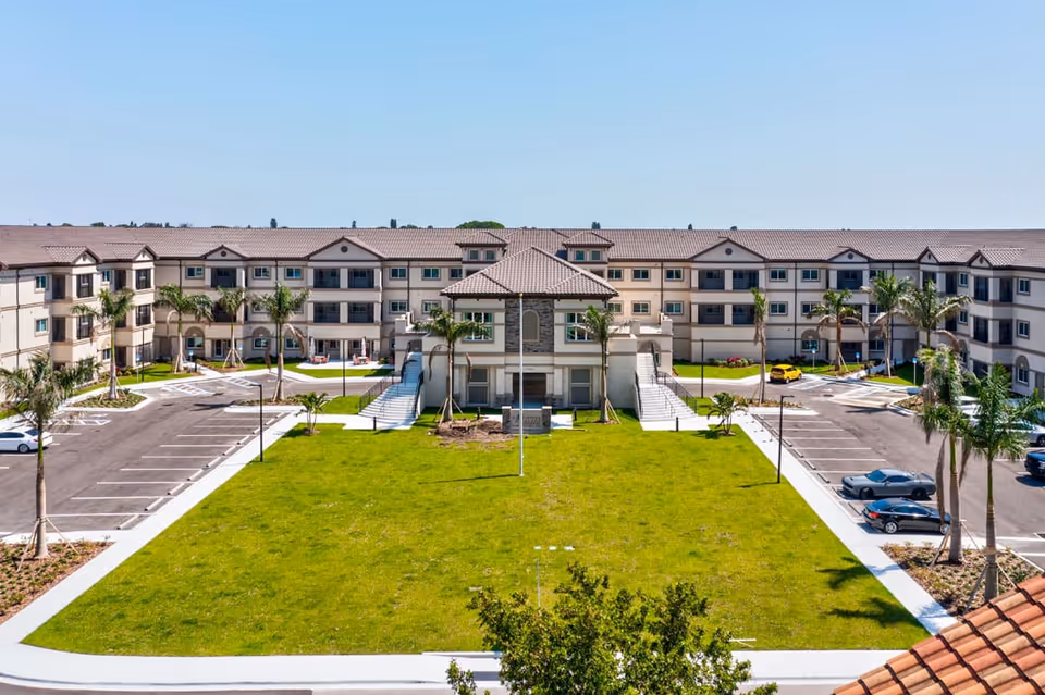 Exterior view of Discovery Village At Sarasota Bay, showing a large U-shaped three-story building with beige walls and a tiled roof. In front of the building is a spacious green lawn with palm trees and a central flagpole, flanked by parking lots on both sides with several parked cars.