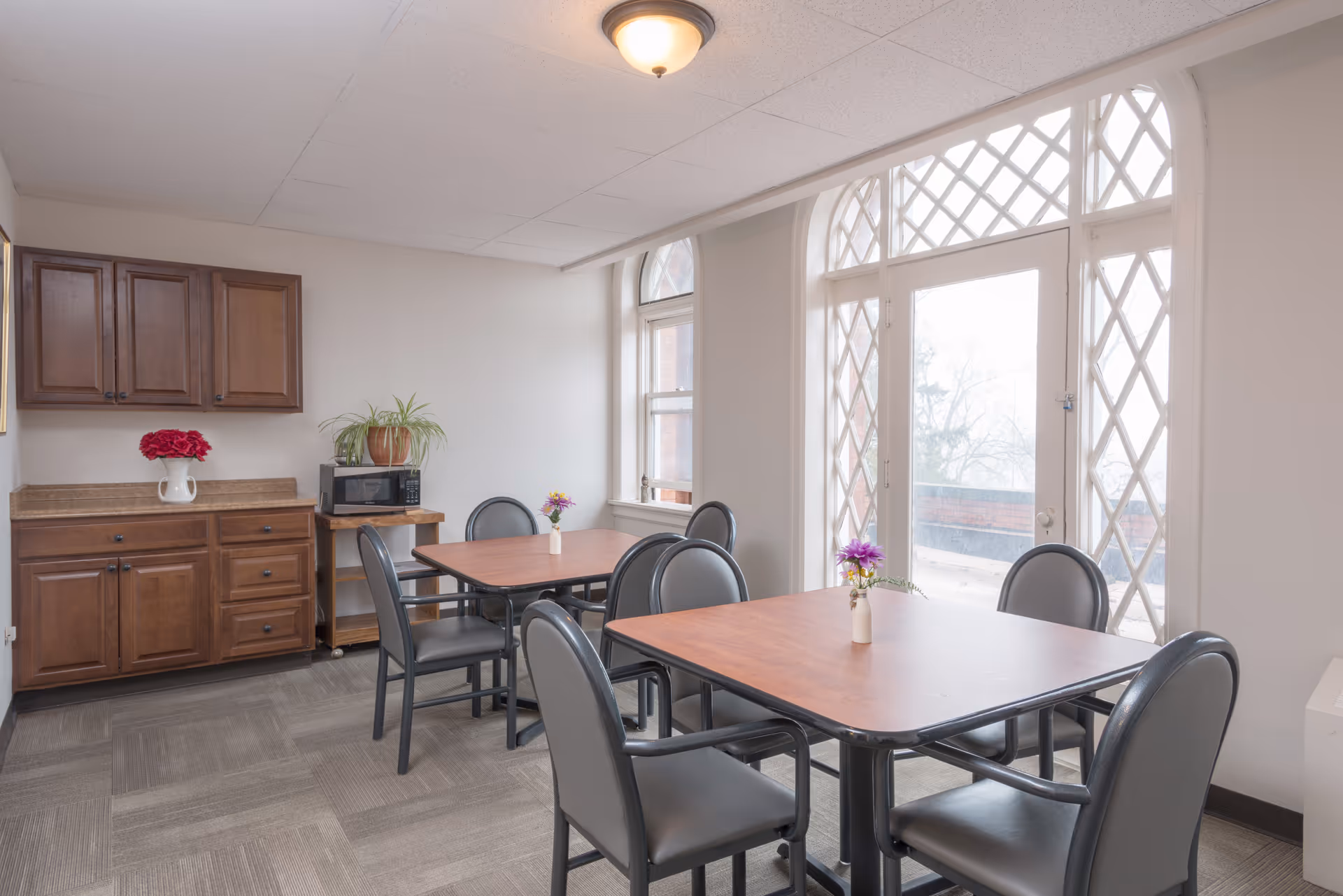 A bright dining area with two wooden tables, each surrounded by four gray cushioned chairs. The room has large windows with decorative lattice patterns allowing natural light to fill the space. There are wooden cabinets on one wall, a microwave on a small wooden cart, and small vases with flowers on each table.