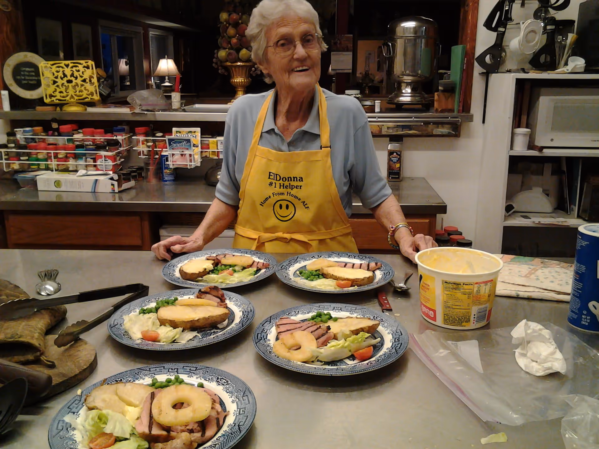 An elderly woman wearing glasses and a yellow apron that reads 'ElDonna #1 Helper Home From Home ALS' stands behind a kitchen counter with five plates of food. Each plate contains a slice of ham with a pineapple ring, a baked potato with butter, peas, lettuce, and a tomato slice. The kitchen has various utensils, spice racks, and appliances visible in the background.