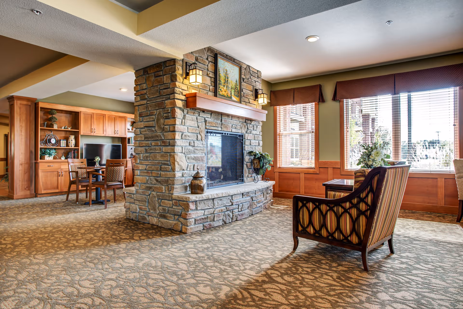 A cozy senior living common area featuring a large stone fireplace in the center, with a framed painting above it. To the right, there is a striped armchair and a small table near large windows with wooden blinds and valances, allowing natural light to fill the room. In the background, there is a wooden cabinet with a TV and a small dining table with chairs. The room has patterned carpet and warm, inviting decor.