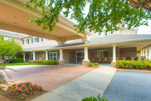 Covered porte-cochère entrance to a multi-story senior living building with columns, outdoor seating, and landscaped shrubs.