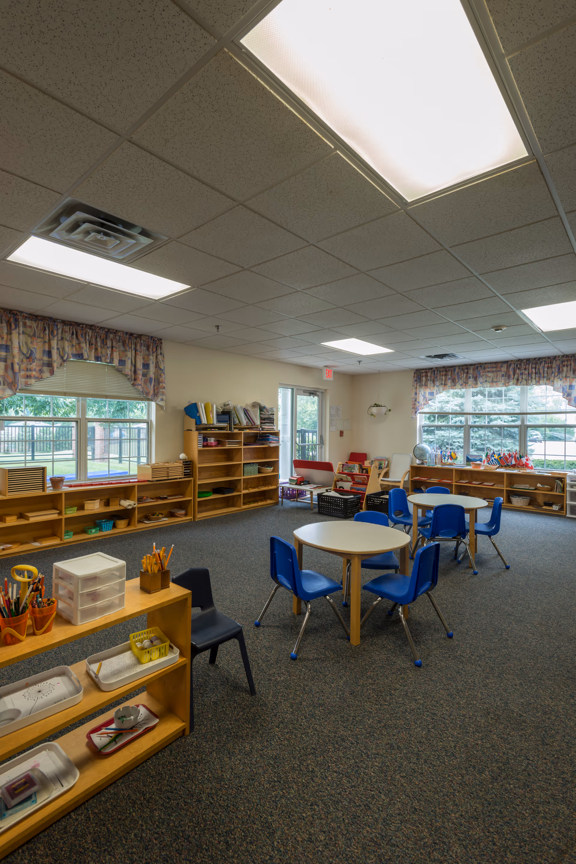 A spacious, well-lit room with carpeted floor featuring small round tables surrounded by blue chairs. Wooden shelves line the walls, holding various educational materials and supplies. Large windows with patterned valances allow natural light to enter, and there are additional seating areas with red chairs near the back. The ceiling has fluorescent lighting panels.