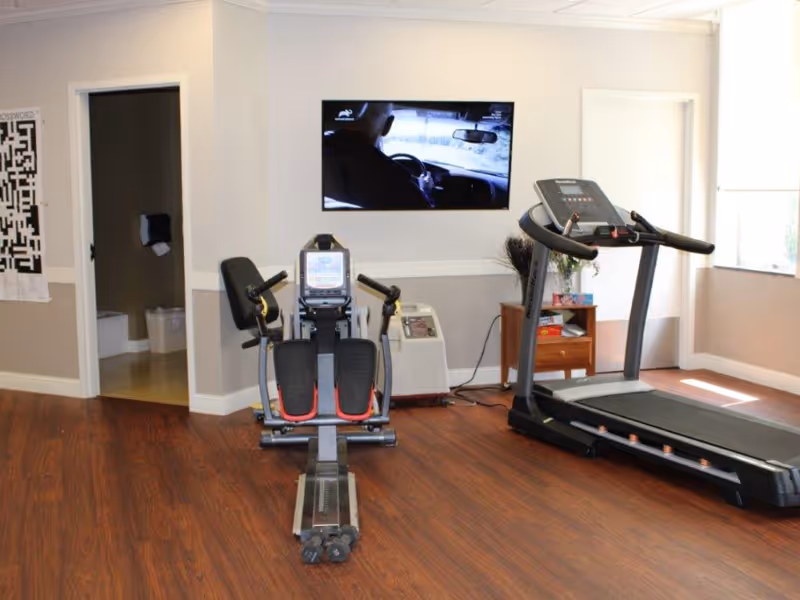 A small exercise room with wooden flooring featuring a rowing machine and a treadmill. A flat-screen TV is mounted on the wall showing a person driving a car. There is a small wooden side table with some items on it and two doorways leading to other rooms.