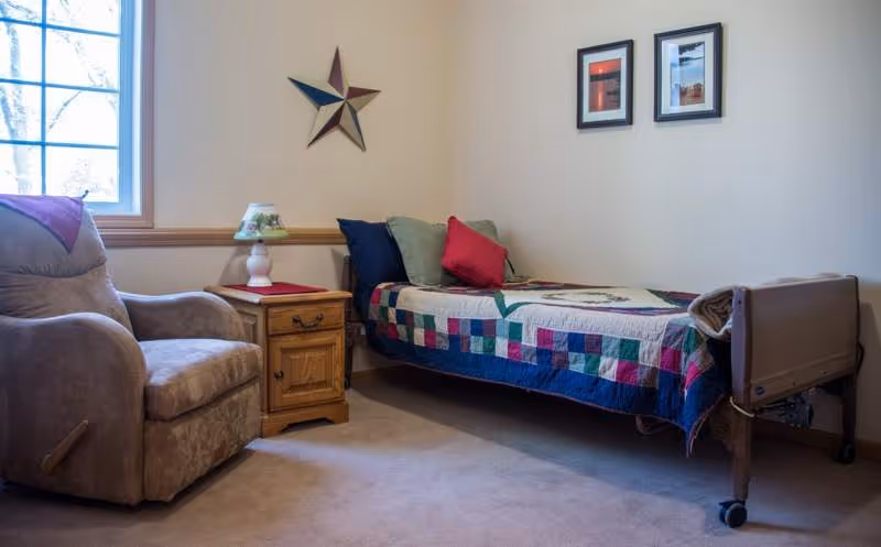 A simple senior living bedroom with a single bed covered by a colorful quilt, a recliner, a wooden nightstand with a lamp, and framed wall decor.