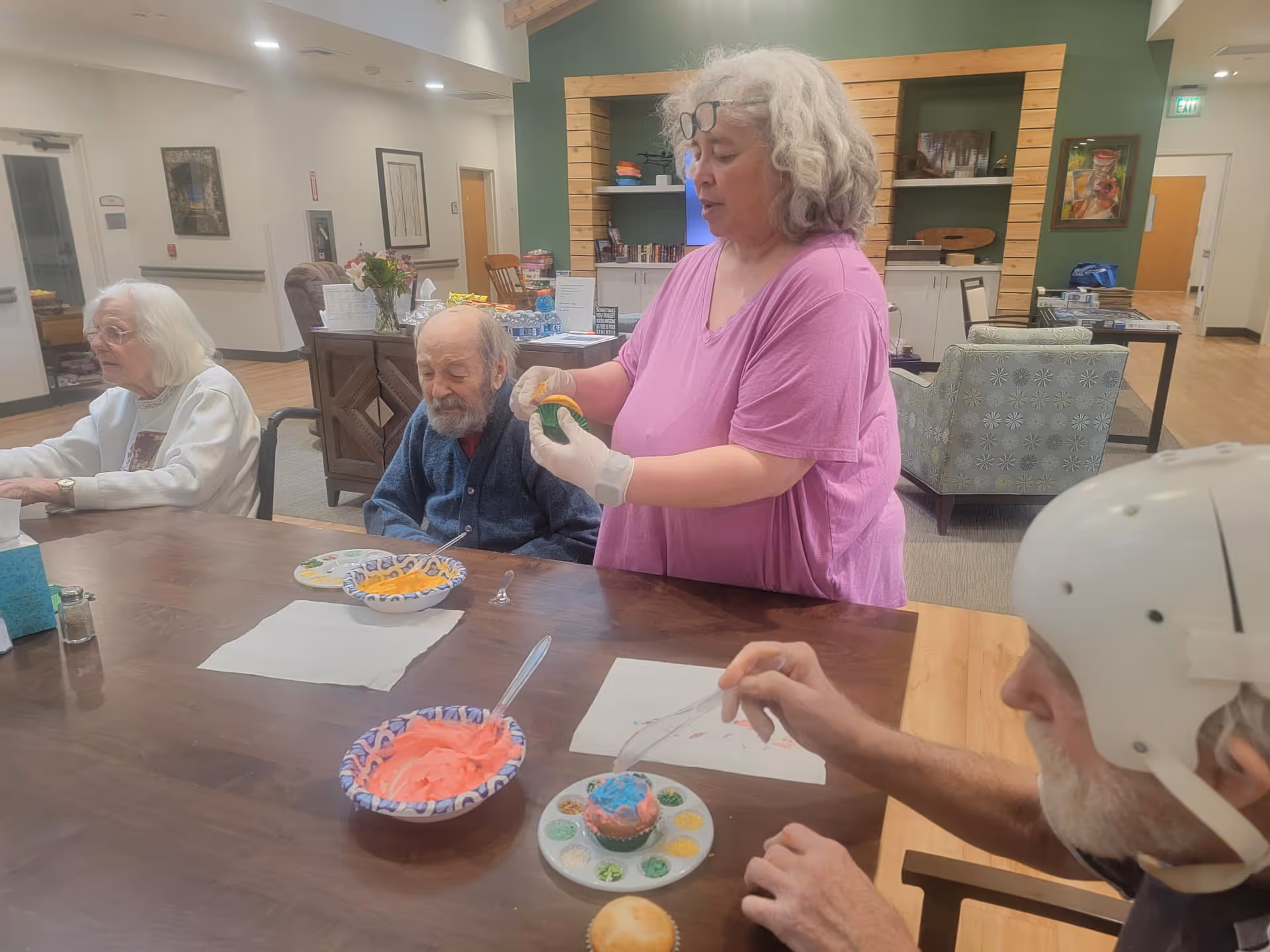A group of elderly people and a caregiver gathered around a table in a common area. The caregiver, wearing a pink shirt and gloves, is holding a decorated cupcake. There are bowls of colorful frosting and a decorated cupcake on the table. The room has comfortable chairs, shelves, and a green accent wall in the background.