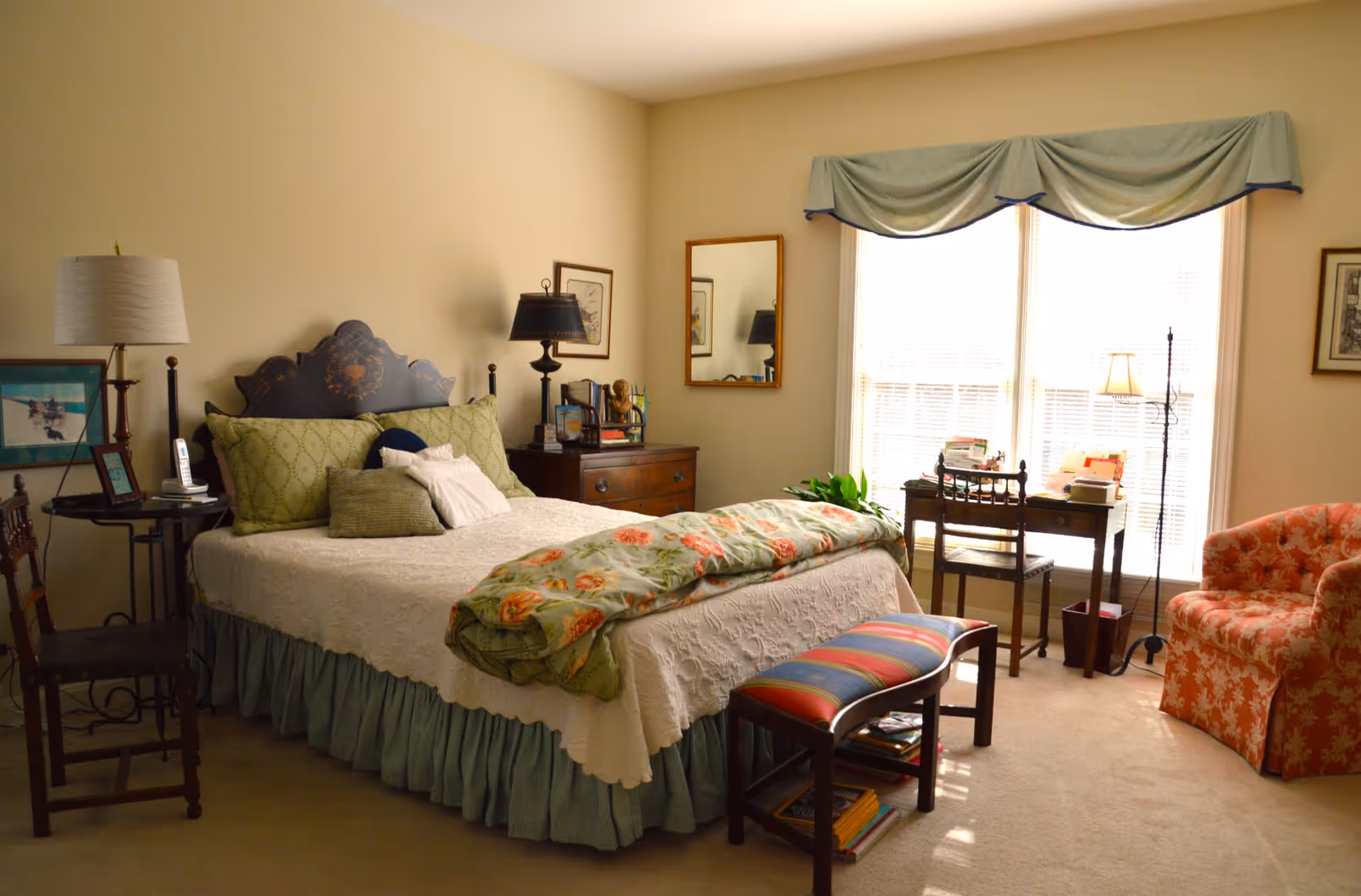A cozy bedroom with a bed covered in a white quilt and green pillows, a floral blanket folded at the foot. There is a wooden bench with a colorful striped cushion in front of the bed. To the left, there is a side table with a lamp and a chair. On the right side, a wooden desk with a chair is placed in front of large windows with blinds and a green valance. A coral-colored armchair with a floral pattern is near the desk. The room has beige walls and carpeted floor.