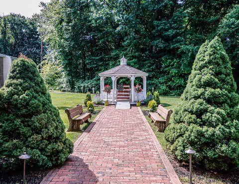 A brick pathway leads to a white gazebo surrounded by green trees and shrubs. There are wooden benches on either side of the pathway and small garden lights along the edges. The scene is outdoors in a well-maintained garden area.