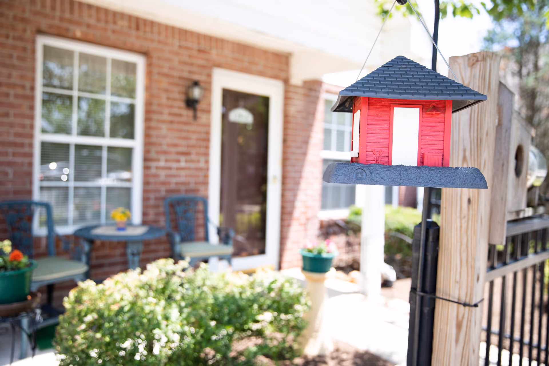 A small red birdhouse hanging from a wooden post in the foreground with a brick building, a glass door, windows, and a small patio area with two chairs and a table in the background.
