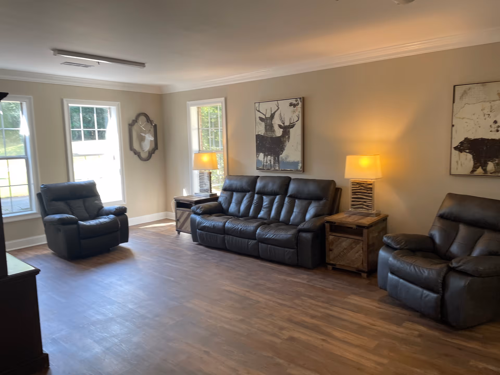 A living room with three black leather recliners, including a three-seat sofa and two single chairs. There are two wooden side tables with lamps on either side of the sofa. The walls are beige with two framed animal-themed artworks and a decorative mirror. The room has large windows letting in natural light and wooden flooring.