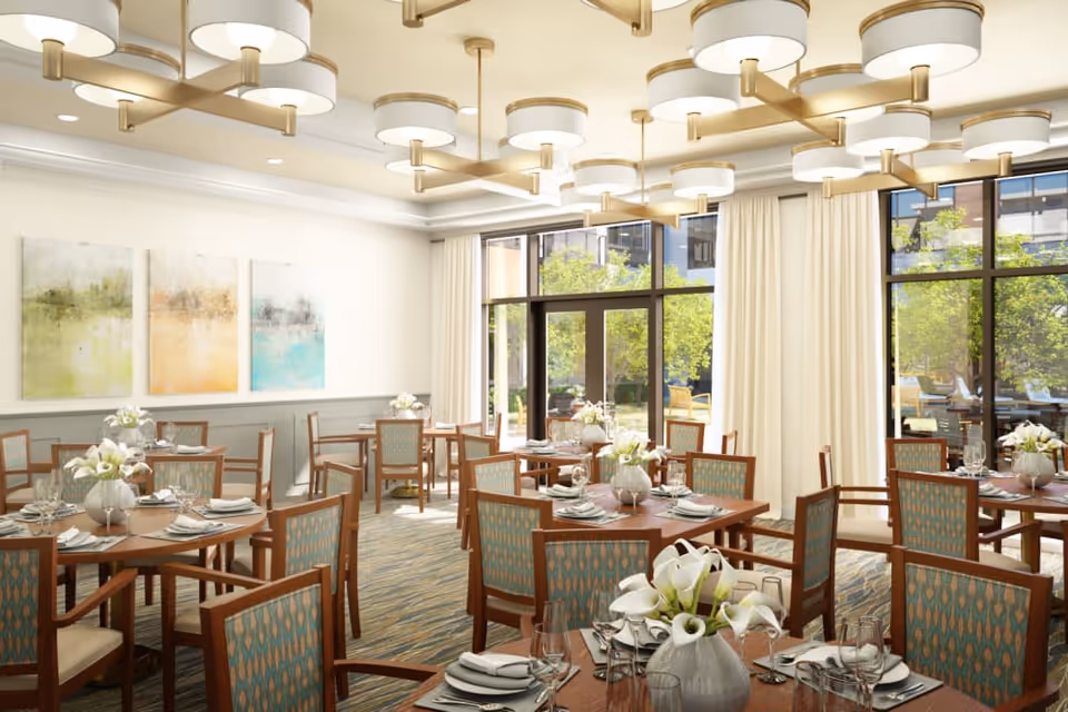 A bright and elegant dining room with multiple wooden tables and chairs arranged neatly. Each table is set with white plates, napkins, glassware, and a vase of white flowers. Large windows with cream-colored curtains allow natural light to fill the room, and abstract paintings hang on the wall. Modern circular ceiling lights provide additional illumination.