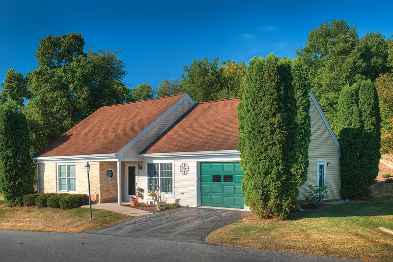 Single-story house with a red roof, beige siding, and a green garage door surrounded by tall green trees and bushes under a clear blue sky.