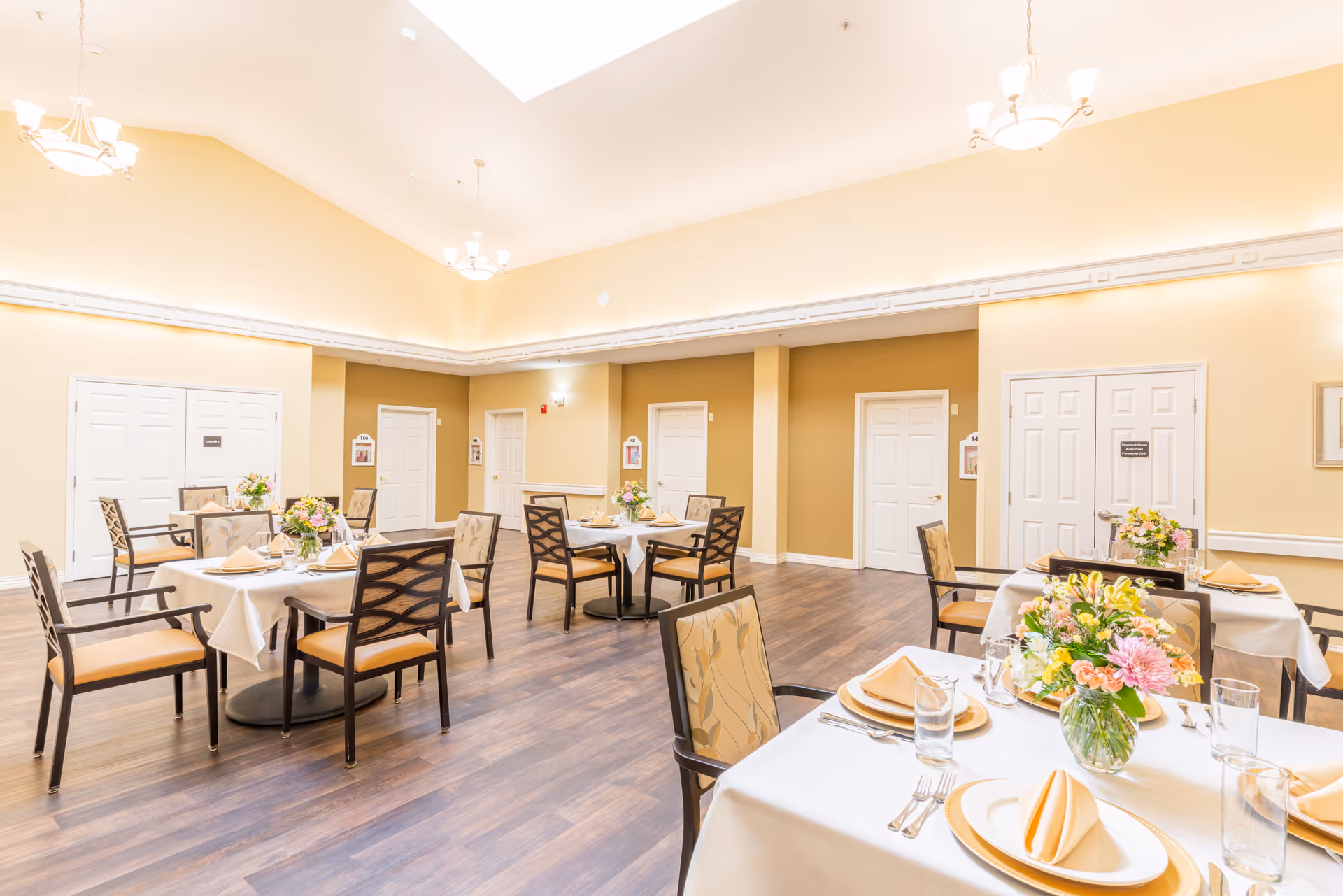 Well-lit communal dining room with multiple tables set with plates, napkins, glasses, and floral centerpieces.