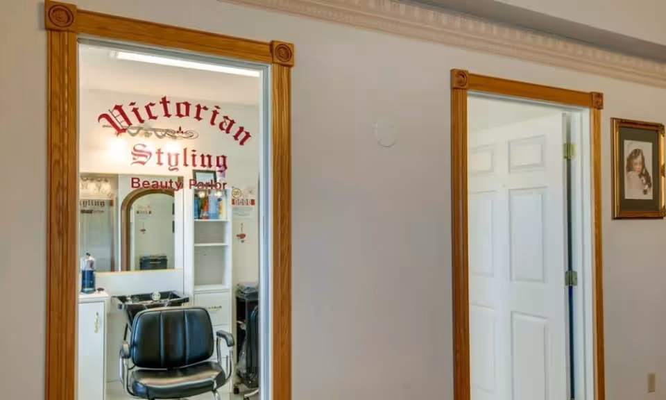 Interior view of a Victorian Styling Beauty Parlor with a black salon chair in front of a mirror and shelves with hair products. The room has wooden door frames and a white door is visible to the right with a framed picture on the wall.
