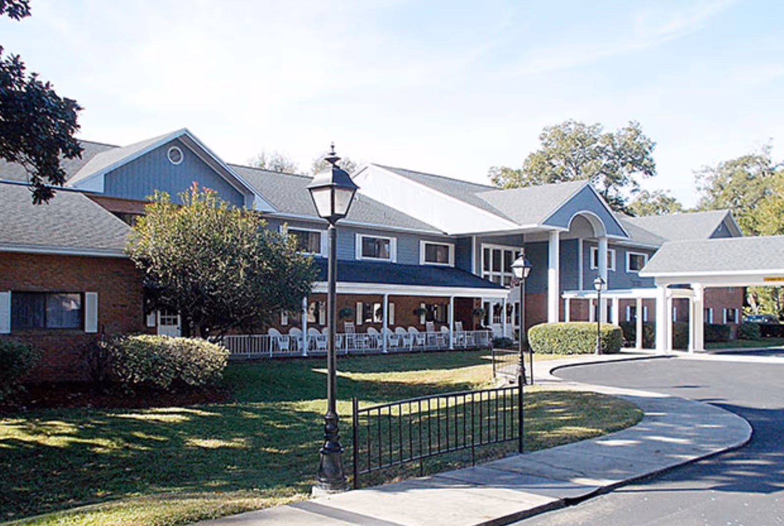 Front exterior of a two-story senior living residence with a covered entrance, porch chairs, lamp posts, and a curved driveway.