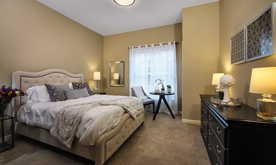 A cozy bedroom with a beige tufted bed frame and white bedding, accented with multiple pillows and a soft throw blanket. There is a small round table with two chairs near a window with sheer white curtains. The room features warm beige walls, a black dresser with lamps and decorative items, and a floor lamp beside the bed.