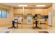 A clean and well-lit kitchen and dining area with light wood cabinets, a white stove, microwave, and refrigerator. There are two small dining tables each with two chairs, and windows with blinds letting in natural light.