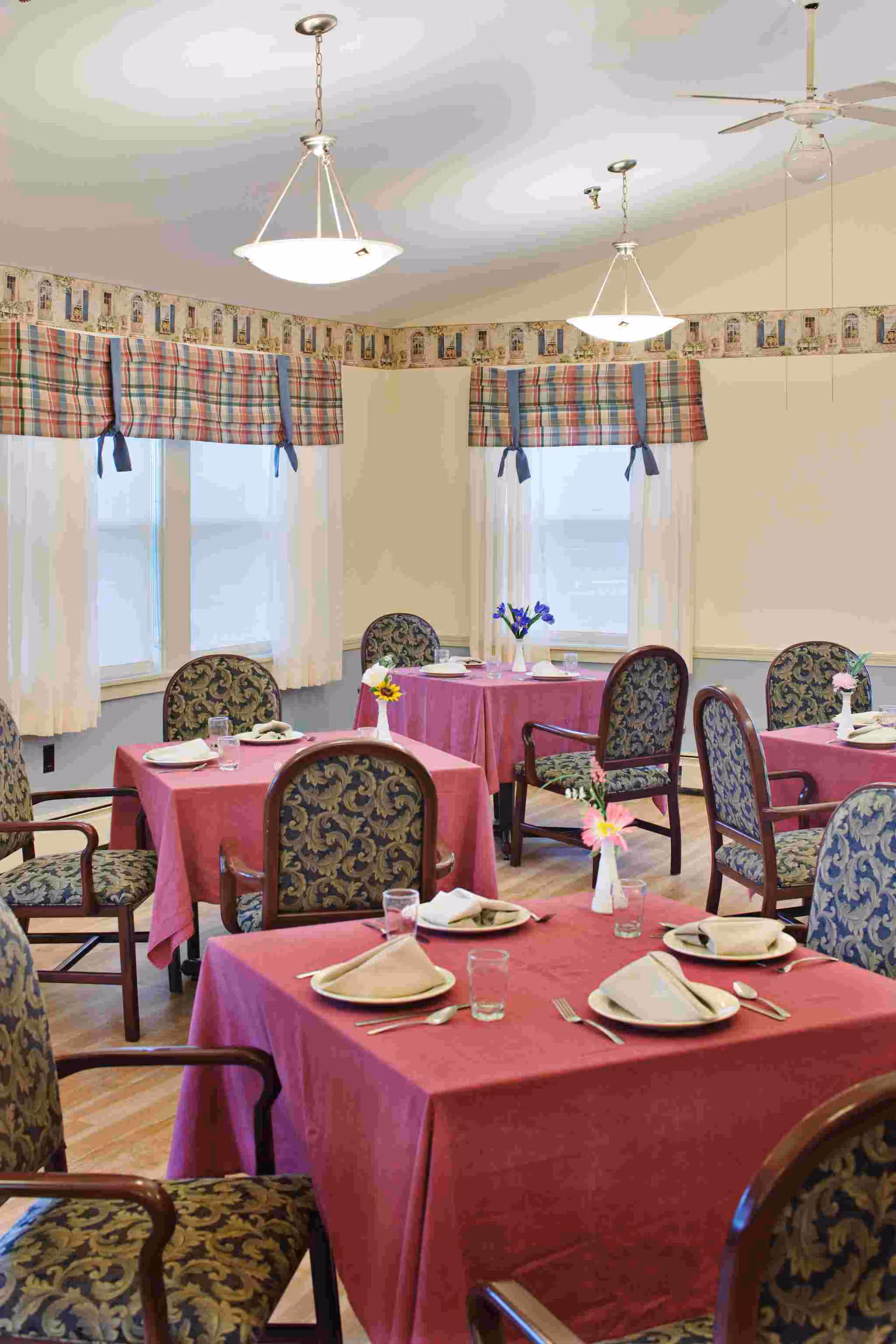 A dining room with several tables covered in red tablecloths, each set with plates, napkins, glasses, and silverware. The chairs have patterned upholstery, and there are small flower vases on each table. The room has windows with plaid valances and cream-colored curtains, and two ceiling light fixtures hang from the ceiling.