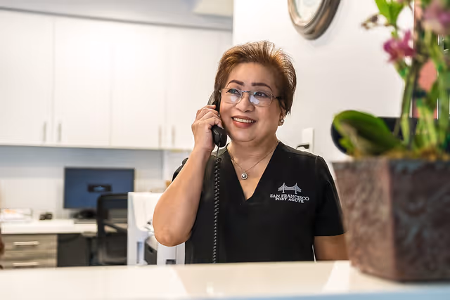 A smiling woman wearing glasses and a black shirt with the San Francisco Post Acute logo is talking on a corded phone behind a reception desk in a bright office setting with white cabinets and a clock on the wall.
