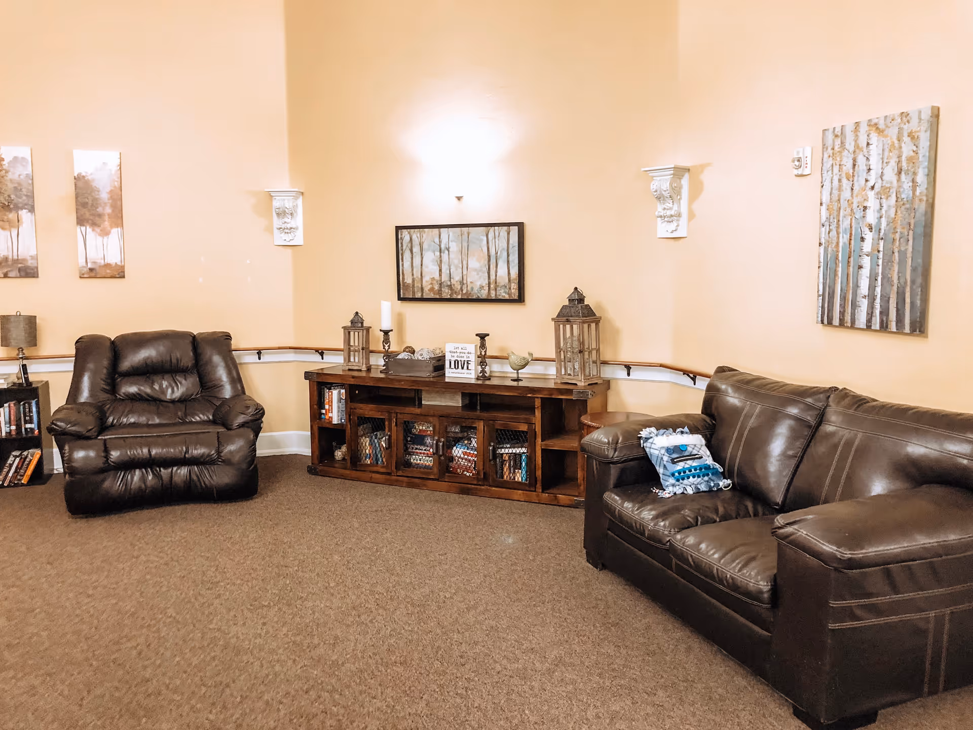A cozy living room with beige walls and carpeted floor. The room features a dark brown leather armchair on the left and a matching leather sofa with a decorative pillow on the right. Between them is a wooden cabinet with books and decorative items on top, including lanterns, candles, and a small sign. The walls are decorated with framed paintings of trees and nature scenes, and there are two white ornamental wall brackets with lights above the cabinet.