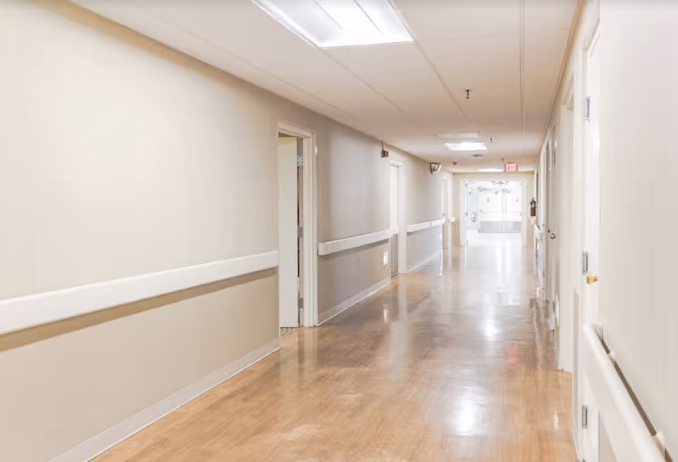 Empty, well-lit nursing facility hallway with handrails, doors along both sides, and a polished floor.