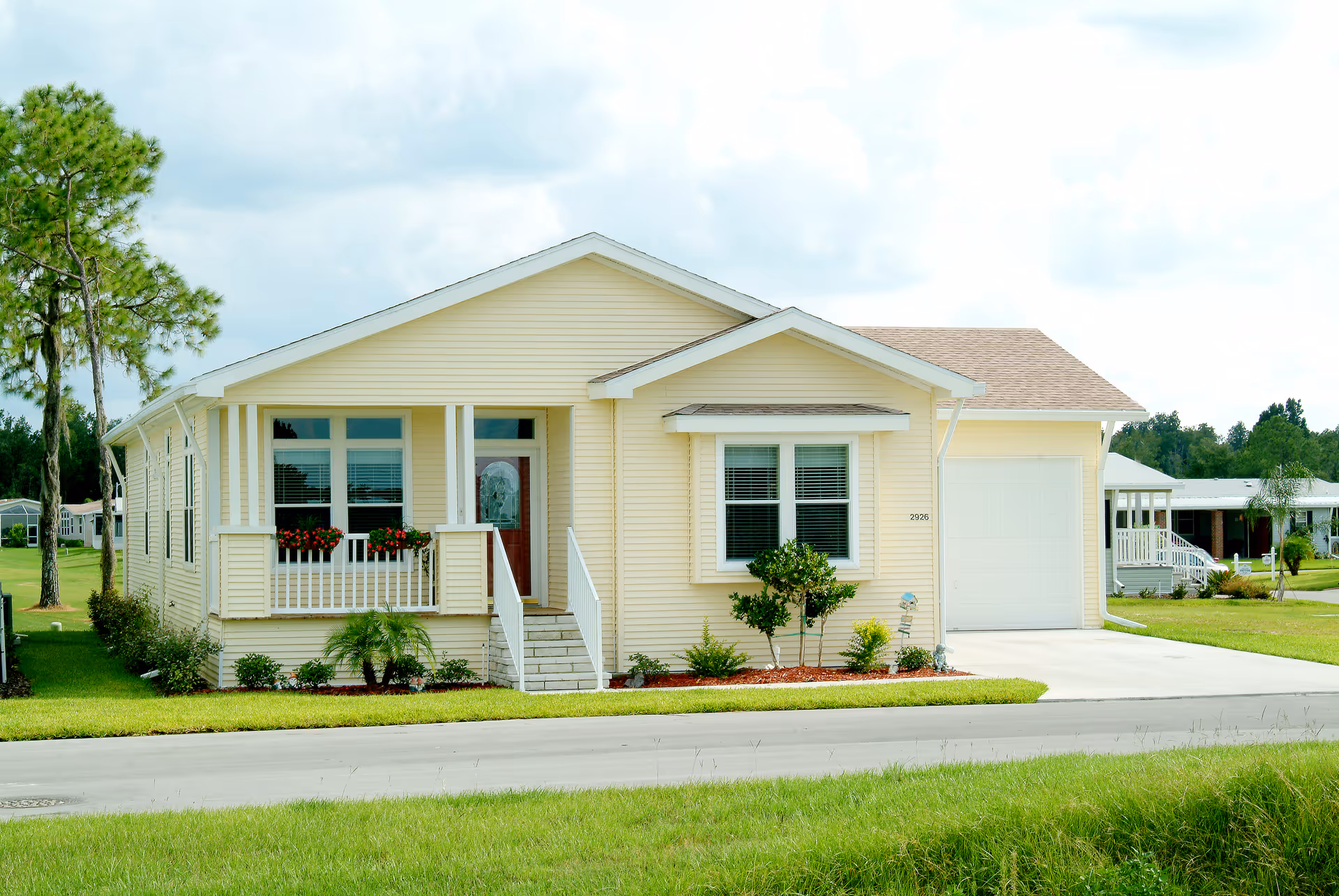 A single-story yellow house with white trim, a small front porch with flower boxes, a front door with glass panel, and an attached garage. The house is surrounded by a well-maintained lawn and landscaping, with a tree on the left side and other houses visible in the background under a partly cloudy sky.