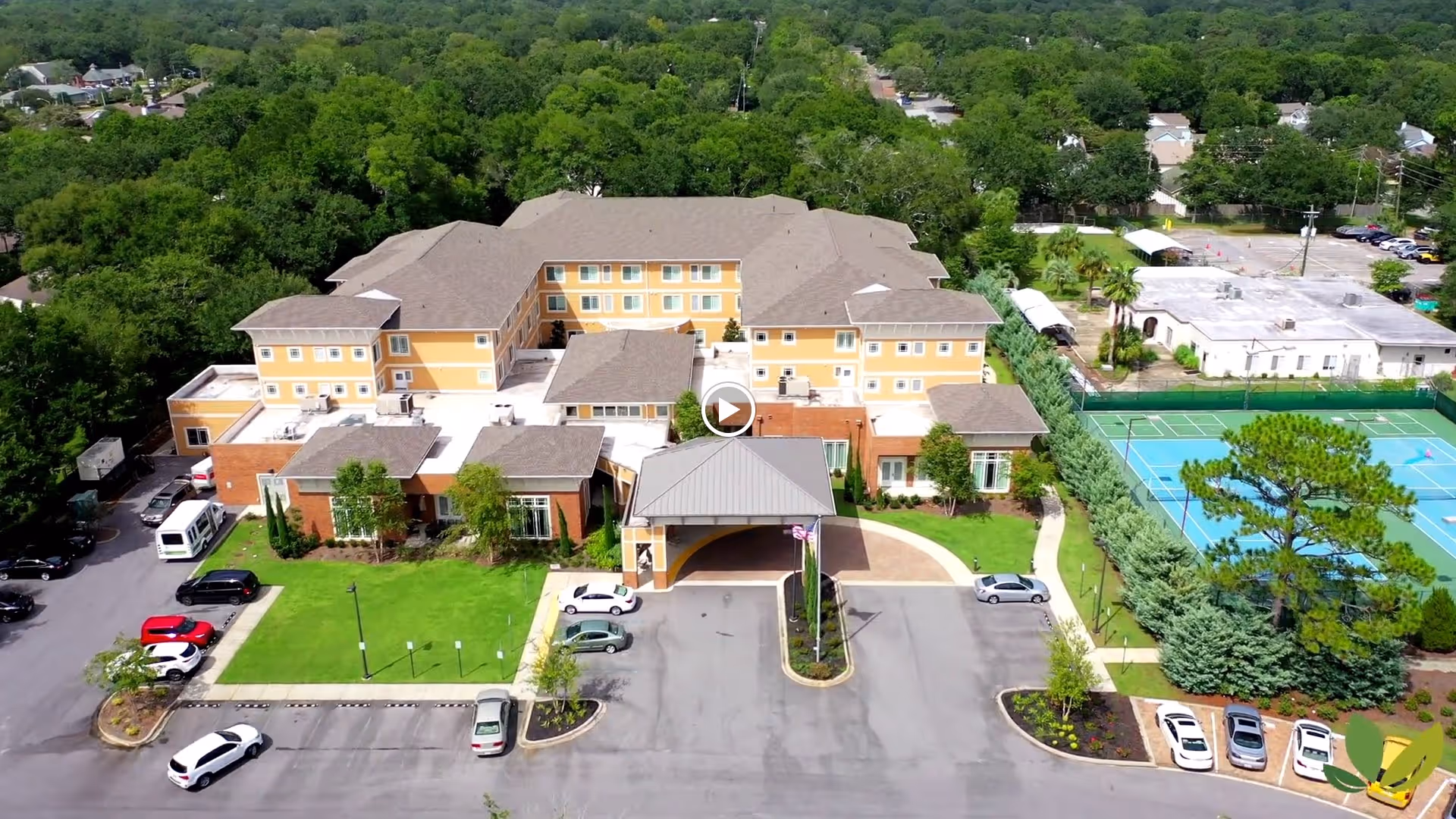 Aerial view of Summer Vista Assisted Living facility showing a large multi-story building with a covered entrance, surrounded by green lawns, trees, parked cars, and a tennis court on the right side.