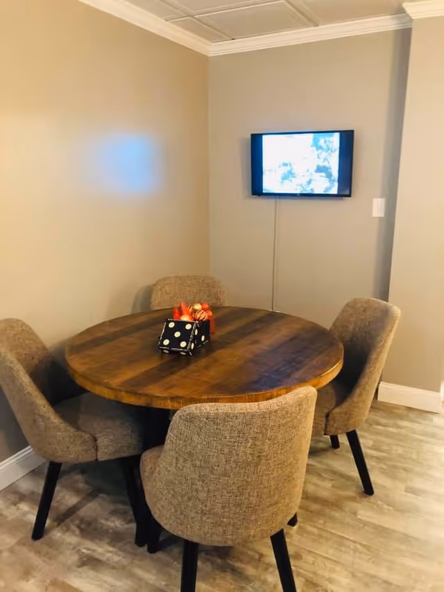 A small dining area with a round wooden table surrounded by four upholstered chairs. On the table, there is a decorative centerpiece with a black and white polka dot box and some red and orange decorative items. A flat-screen TV is mounted on the beige wall in the background.