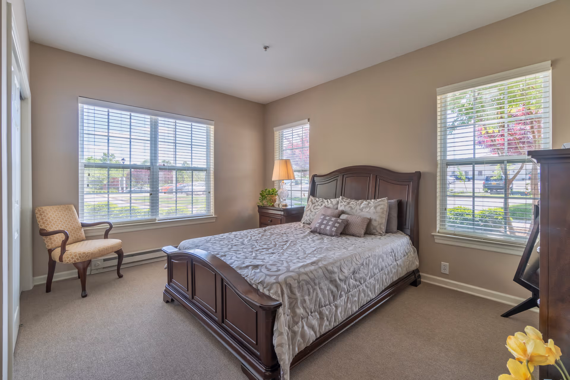 A bright bedroom with beige walls and carpet, featuring a wooden bed with a patterned comforter and multiple pillows. There are three large windows with white blinds letting in natural light. A wooden nightstand with a lamp and a plant is beside the bed, and an upholstered chair is placed near one window. A wooden dresser is partially visible on the right side of the room.