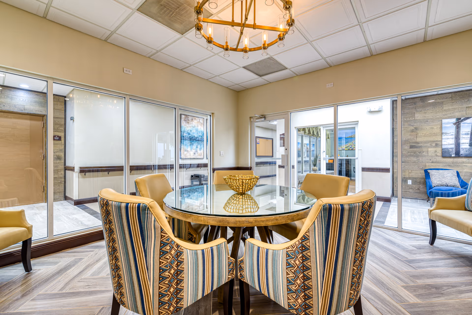 A bright and modern common area in a senior living facility featuring a round glass-top table surrounded by six upholstered chairs with patterned and solid yellow fabric. The room has large windows and glass doors allowing natural light to enter, with a chandelier hanging from the ceiling. The flooring is a light wood pattern, and there are artworks on the walls visible through the glass.