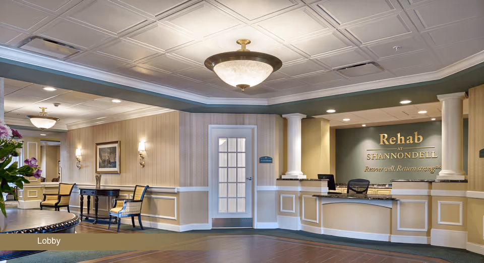 Lobby area of Rehab At Shannondell facility featuring a reception desk with a sign behind it that reads 'Rehab at Shannondell Recover well. Return strong.' The lobby has beige walls with white trim, a round table with flowers, chairs, a painting on the wall, and ceiling light fixtures.