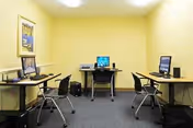 Small computer room with three desks arranged along the walls, each desk equipped with a desktop computer and chair. The walls are painted yellow, and there is a framed picture hanging on the left wall. The floor is carpeted in dark gray.