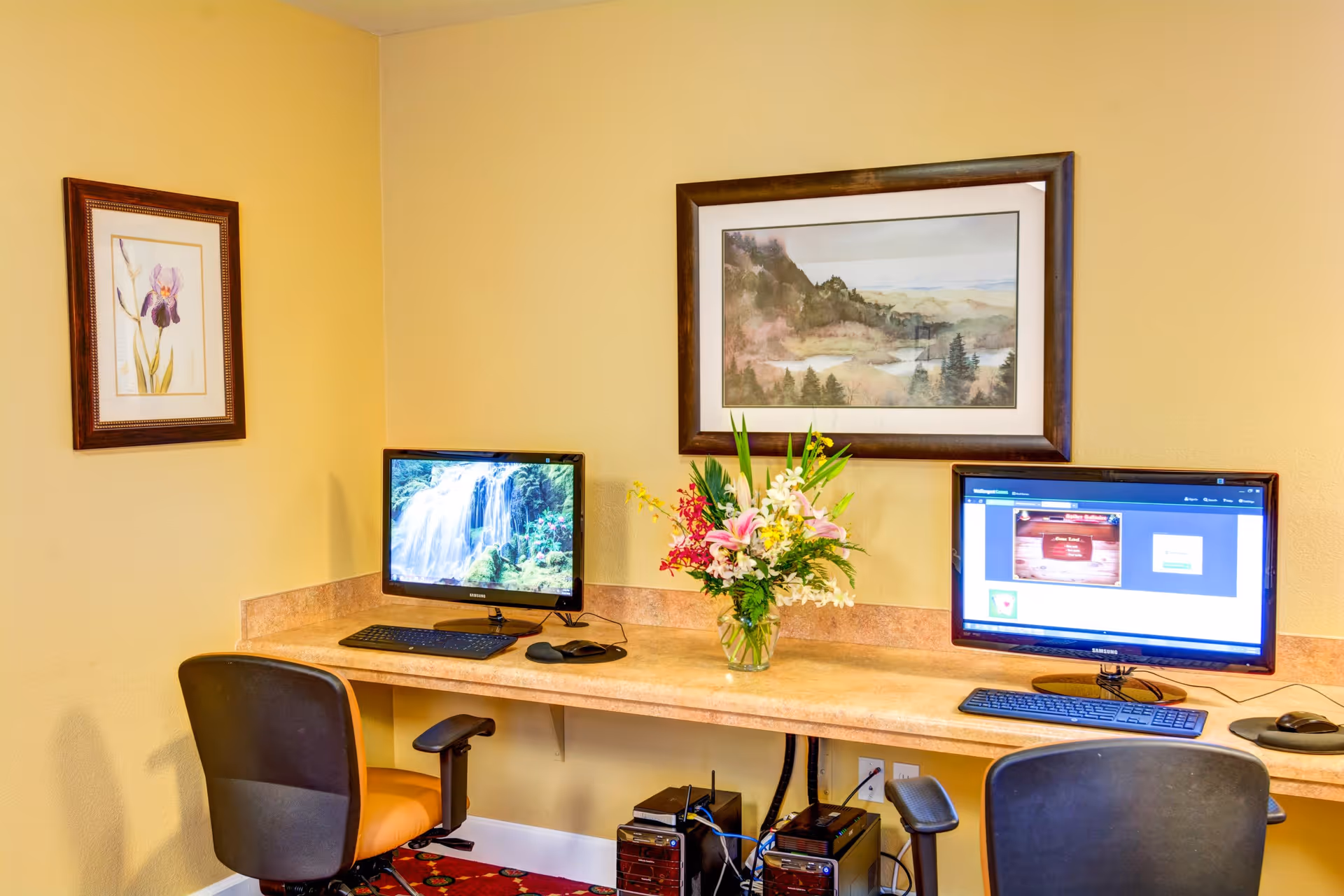 A small computer workstation area with two desktop computers on a long counter against a yellow wall. Each computer has a keyboard and mouse, and there are two office chairs in front of the counter. A vase with a colorful bouquet of flowers is placed between the two monitors. The wall is decorated with two framed pictures, one of a flower and the other of a landscape.