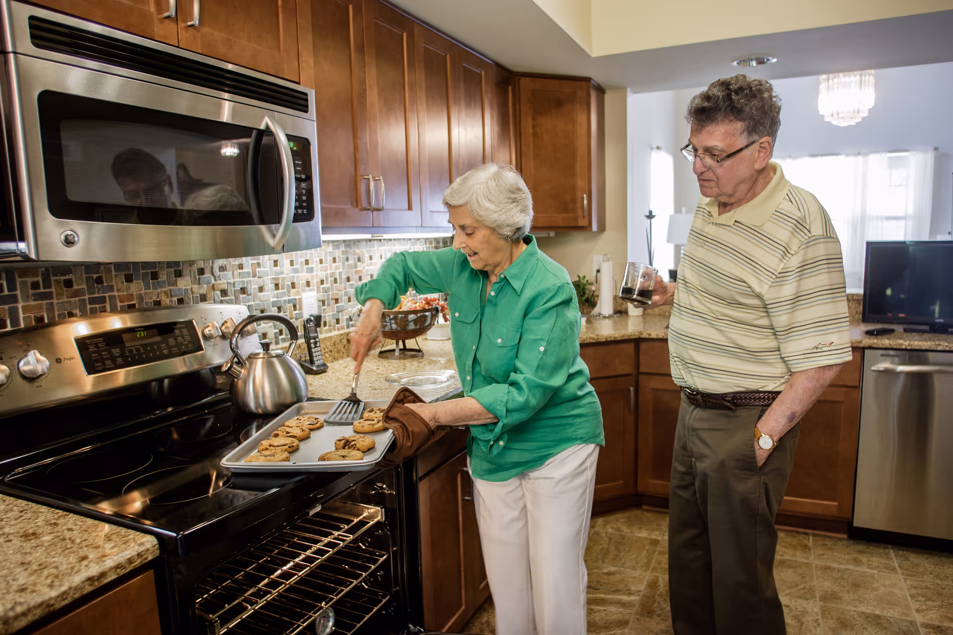 An elderly woman wearing a green shirt and white pants is taking a tray of freshly baked cookies out of an oven in a kitchen. An elderly man in a striped polo shirt and brown pants stands nearby holding a glass, watching her. The kitchen has wooden cabinets, a tiled backsplash, and granite countertops.