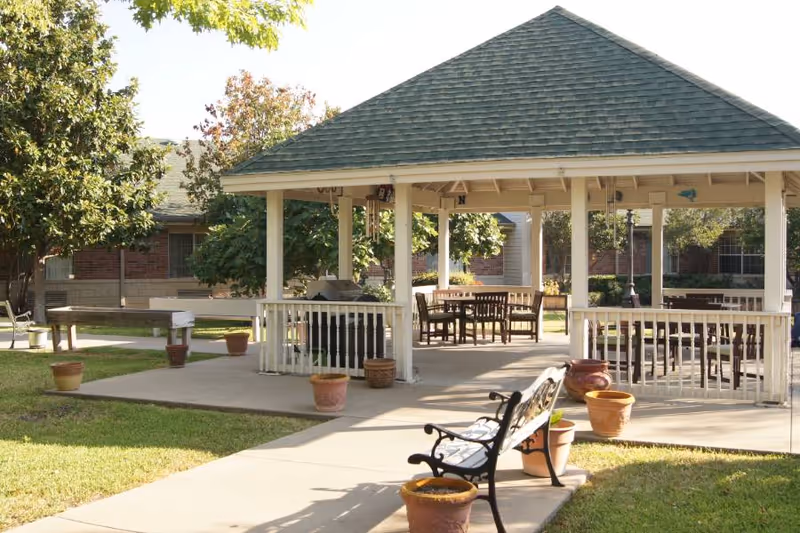 Outdoor covered pavilion with a green shingled roof, surrounded by trees and grass. Several wooden tables and chairs are placed under the pavilion. A black metal bench and multiple empty terracotta pots are positioned on the concrete walkway leading to the pavilion. A brick building is visible in the background.