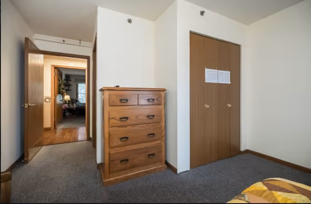 A simple bedroom interior with a wooden chest of drawers against a white wall, a closed wooden closet with two doors, and an open doorway leading to another room with wooden flooring and furniture visible.