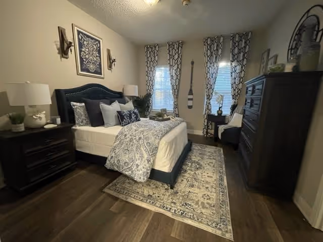 A neatly arranged bedroom with a large bed featuring a dark blue upholstered headboard, white and blue patterned bedding, and multiple pillows. The room has dark wooden flooring with a decorative area rug under the bed. There are two windows with patterned curtains, a dark wooden dresser, a small side table with a lamp, and wall decorations including a framed artwork and a decorative paddle.