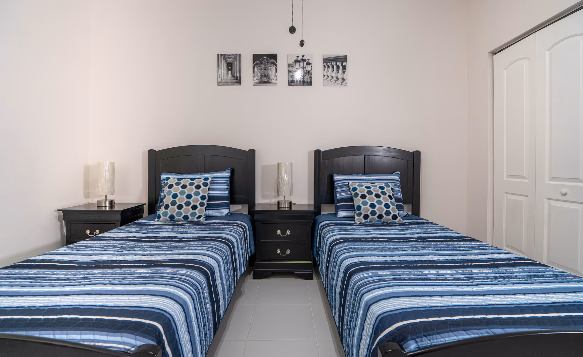 A bedroom with two single beds side by side, each with a blue striped bedspread and a decorative pillow. Between the beds is a dark wooden nightstand with a lamp on top. Above the beds are four black and white framed pictures on the wall. To the right is a white closet with sliding doors.