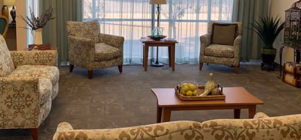 A cozy living room area with patterned upholstered sofas and armchairs arranged around a wooden coffee table. The coffee table has a decorative tray with a glass container of lemons and other decorative items. Behind the seating area is a large window with sheer curtains allowing natural light to fill the room. There are plants and decorative items placed around the room, creating a warm and inviting atmosphere.