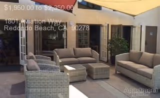 Outdoor patio area with two sofas, two armchairs, and two small tables arranged under a large beige canopy. The seating furniture is made of wicker with cushions. The patio is adjacent to a building with open French doors and potted plants.
