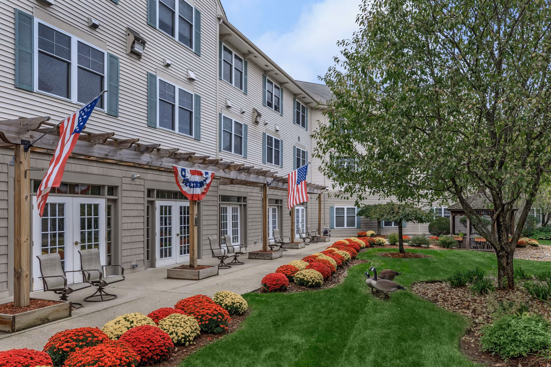 Outdoor garden area of a senior living facility with a row of red and yellow flowers along a concrete walkway. There are several American flags and patriotic bunting hanging from a wooden pergola attached to the building. The building has multiple windows and several chairs are placed along the walkway. A tree and green grass are visible on the right side of the image.