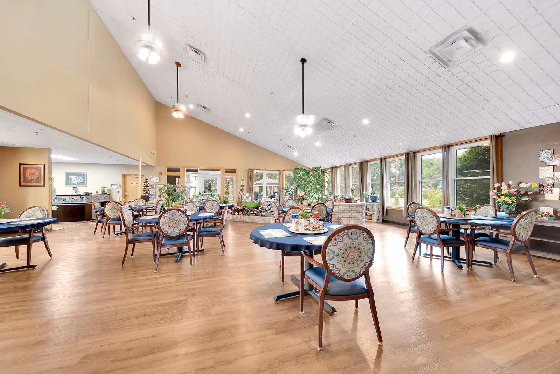 A spacious dining room with multiple round tables covered with blue tablecloths and patterned chairs. The room has large windows letting in natural light, plants, and ceiling fans with lights. The floor is wooden, and there are decorative items and artwork on the walls.