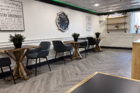Bright communal dining area with small wooden tables, gray chairs, wall clock and potted plants.