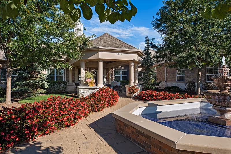 Entrance of a senior living building with a columned portico, landscaped walkway, red flowers, and a fountain.