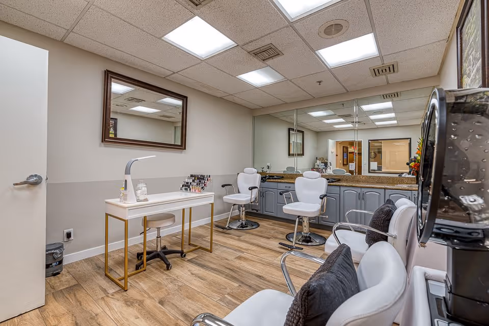 Interior view of a salon area in a senior living facility with white salon chairs, a manicure table with nail polish bottles, a large mirror on the wall, and wooden flooring.