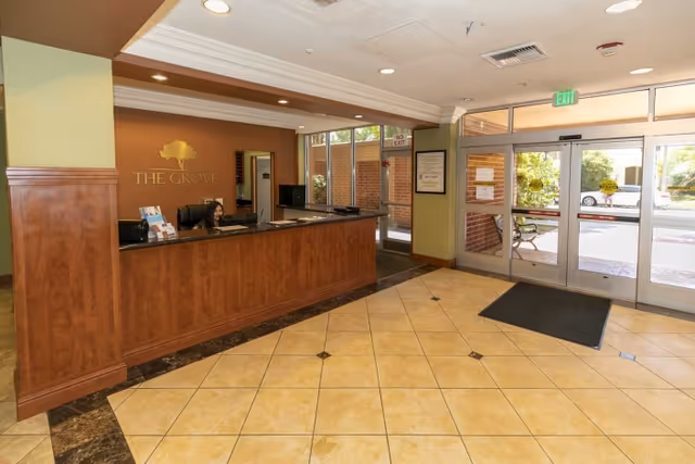 Reception lobby with a wooden front desk, tiled floor, and glass entrance doors with 'The Grove' logo on the wall.