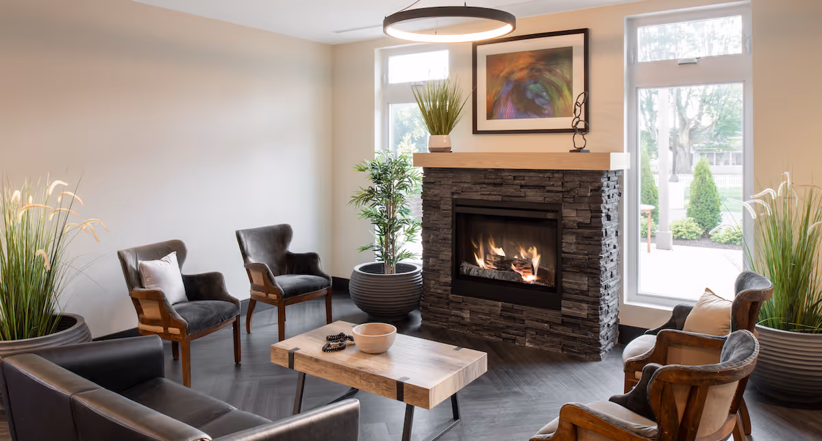 A cozy living room with a modern stone fireplace with a fire burning inside. The room features a wooden coffee table, several armchairs with cushions, large potted plants, and two tall windows letting in natural light. A colorful abstract painting hangs above the fireplace.